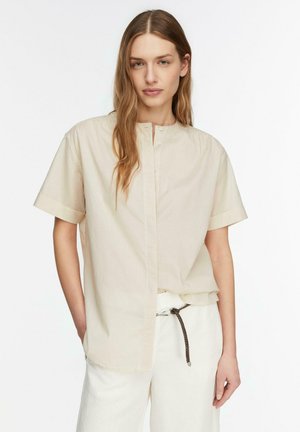 Frau mit langen Haaren, die ein beiges, kurzärmliges, geknöpftes Hemd und weiße Hose mit einem dünnen braunen Gürtel trägt, steht vor einem einfarbigen Hintergrund.