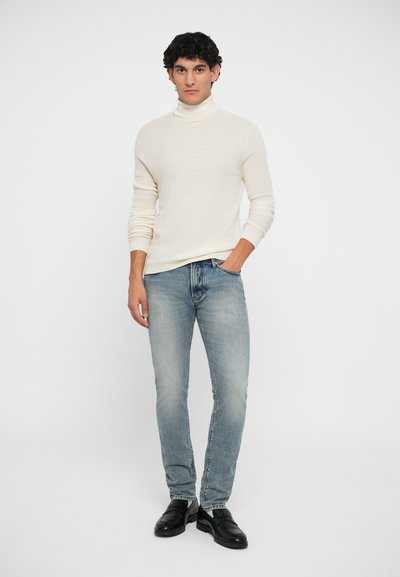 Fehér, kötött garbós pulóver világoskék, slim-fit farmernadrággal és fekete cipővel párosítva. A pulóver textúrált mintával rendelkezik.
