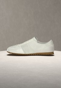 Basket blanche décontractée avec design à lacets et semelle en caoutchouc marron dentelée sur fond neutre.