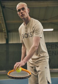 Camiseta deportiva color beige claro con el logotipo "BOSS" en rayas oscuras, acompañada de pantalones cortos a juego. Sosteniendo una raqueta de tenis naranja y una pelota verde.