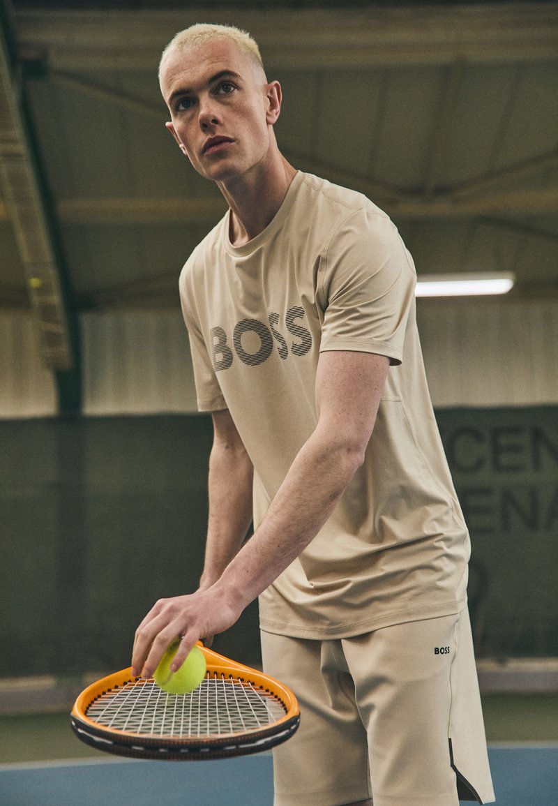 Camiseta deportiva color beige claro con el logotipo "BOSS" en rayas oscuras, acompañada de pantalones cortos a juego. Sosteniendo una raqueta de tenis naranja y una pelota verde.