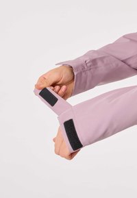 Svetlo vijolična majica z dolgimi rokavi z manšetom, ki ima trak na ježka. Gladek material s prilagojeno prilegajočo obliko. Roke prikazane med nastavljanjem manšete.