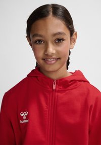 Rote Kapuzenjacke mit voller Zip, ausgestattet mit einem weißen Logo auf der linken Brust. Glattes Material und flache Nähte für zusätzlichen Komfort.