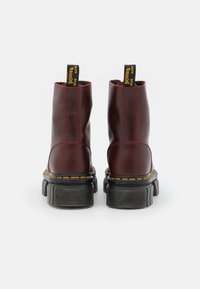 Botas de tobillo de cuero burdeos con una suela de goma negra, costuras amarillas y una pestaña trasera para facilitar el uso. Textura suave, diseño elegante.