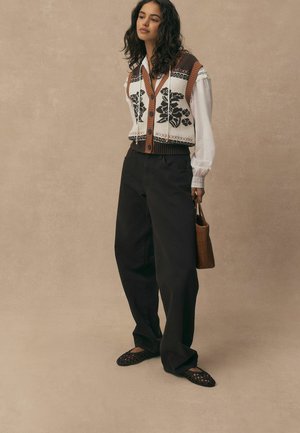 Jauna sieviete baltā blūzē, ar ziedu raksta adītu vestīti, vaļīgām melnām biksēm, austu brūnu somiņu un melnām austām kurpēm uz gaiši brūna fona.