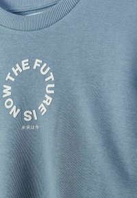 T-shirt bleu à col ras du cou avec un texte circulaire blanc "THE FUTURE IS NOW" et de petits caractères japonais sous le cercle sur la poitrine.