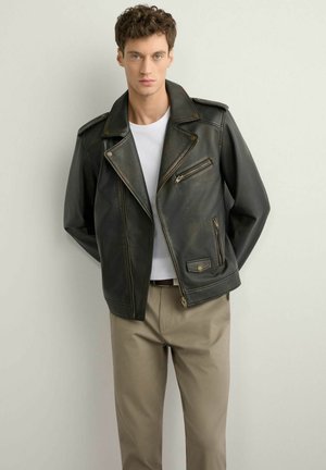 Młody mężczyzna w ciemnej skórzanej kurtce motocyklowej, białej koszulce i beżowych spodniach, stojący na tle jasnej, jednolitej ściany.