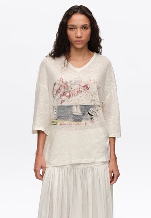 Joven mujer con cabello largo y rizado, vistiendo una camiseta beige holgada con un diseño de velero y una falda blanca plisada, de pie sobre un fondo blanco.