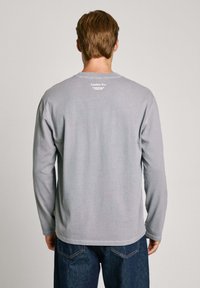Graues Langarm-T-Shirt mit rundem Ausschnitt, mit bedrucktem Branding auf der Rückseite, kombiniert mit blauen Jeans. Glatte Baumwollstruktur.