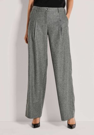 Vrouw draagt grijze krijtgestreepte wijde broek met plooien en zwarte puntige hakken, staand tegen een effen lichte achtergrond.