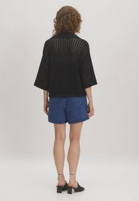 Zwarte gebreide open-top met korte mouwen, met een losse pasvorm en een geribbelde zoom, gecombineerd met blauwe denimshorts en zwarte sleehakken.