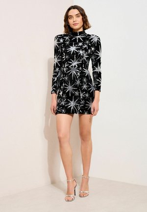 Sieviete, kura valkā melnu samta mini kleitu ar garām piedurknēm un sudraba zvaigžņu sprādziena rakstu, savienotu ar sudraba augstpapēžu sandales, stāvot.