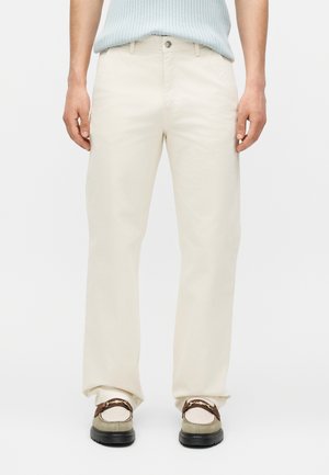 Férfi, aki bézs egyenes szárú nadrágot, világoskék bordázott felsőt hordberakva, és bézs loafert sötétbarna pánttal és arany lánc díszítéssel.