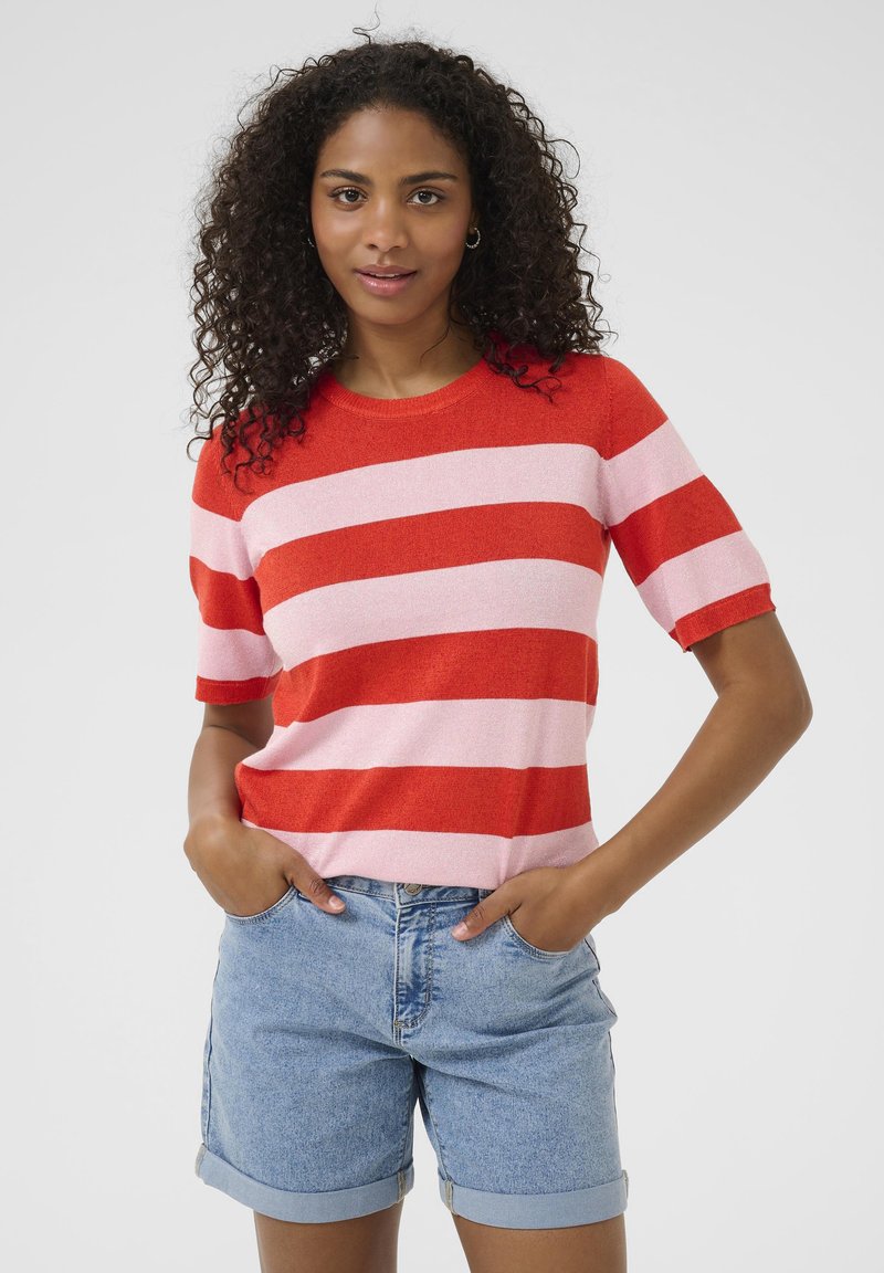 Pull en tricot rayé rouge et rose à manches courtes; porté avec un short en denim bleu clair, les mains dans les poches, tissu texturé.