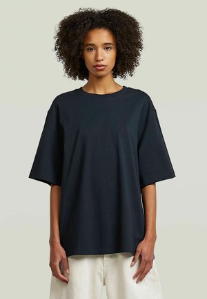 Kobieta z kręconymi włosami, ubrana w luźny czarny, oversize t-shirt i jasne spodnie, stojąca na tle jednolitego neutralnego tła.