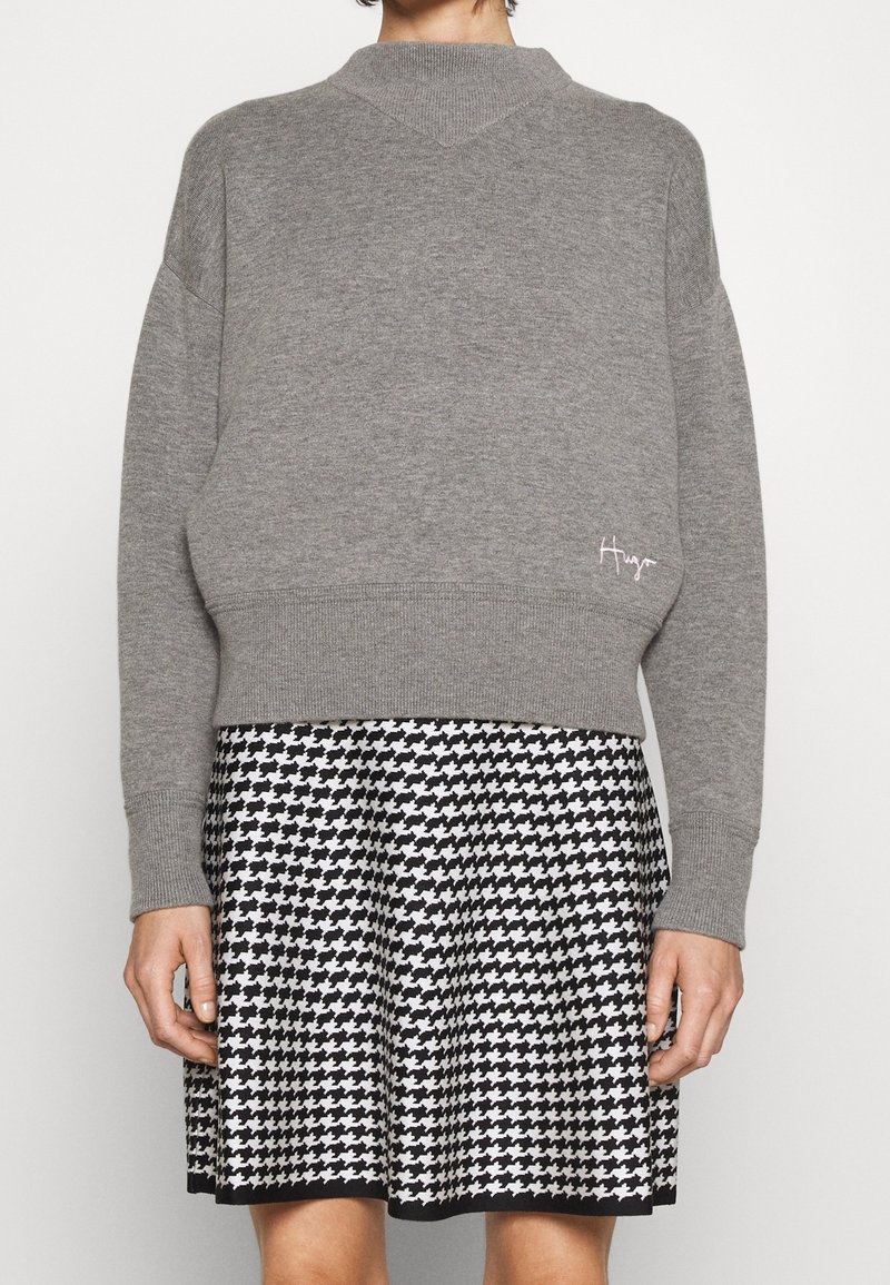 Szary dzianinowy sweter z wysokim dekoltem i prążkowanymi mankietami, w połączeniu z czarno-białą spódnicą w wzór houndstooth, wykończoną na dole.