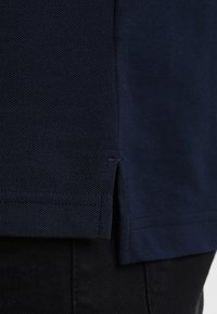 Polo de color navy con una tela texturizada, que cuenta con una abertura lateral para mejorar el ajuste. El dobladillo es recto con detalles de costura bien acabados.