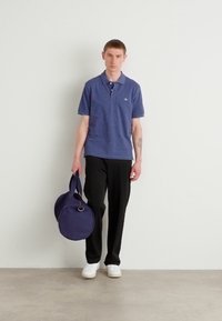 Marineblaues Polo-Shirt mit Kragen, schwarze Hose und weiße Sneakers. Hält eine runde marineblaue Tasche. Gleichmäßige Beleuchtung auf einem neutralen Hintergrund.