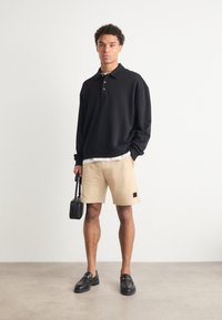 Svart långärmad polotröja, beige shorts, svarta loafers. Modellen håller en liten svart väska. Slätt tyg och avslappnad passform.