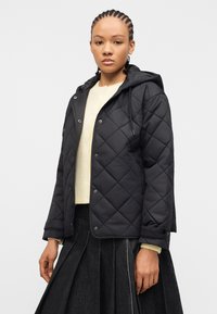 Νεαρή γυναίκα που φοράει μαύρο καπιτονέ μπουφάν με κουκούλα, κρεμ πουκάμισο και πλισέ μαύρη φούστα, στέκεται μπροστά σε ένα απλό ανοιχτό φόντο.