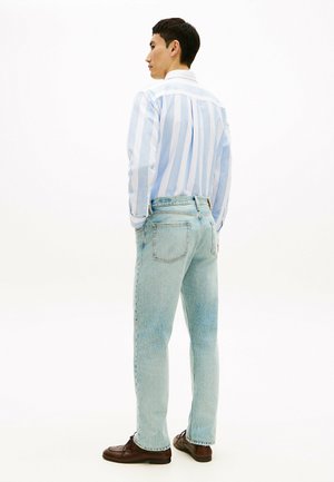 Άνδρας στραμμένος προς τα αριστερά, φορώντας ανοιχτό μπλε και άσπρο ριγέ πουκάμισο, τζιν ανοιχτού χρώματος και καφέ παπούτσια, στέκεται σε απλό άσπρο φόντο.