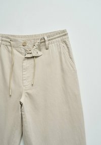 Pantalones casual beige claro con cintura elástica, cierre de botón, cordón, trabillas para cinturón y bolsillos laterales sobre un fondo liso.