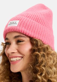 Różowa, prążkowana czapka z dzianiny z wplecioną białą metką z napisem "ROXY". Miękka tekstura z luźnym krojem, zaprojektowana dla ciepła i komfortu.