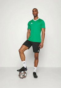Cămașă sportivă verde cu guler alb, pantaloni scurți negri, ținând o minge de fotbal. Prezintă logo-ul Adidas atât pe cămașă, cât și pe pantaloni, cu un design elegant.