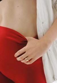 Tela de satén roja se agrupa en la cintura, con una mano agarrando el material. En la mano se lleva un anillo dorado con un diseño ondulado.
