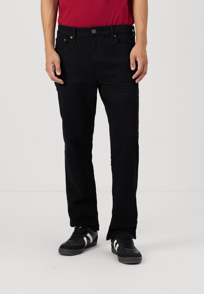 Pantalones vaqueros negros de mezclilla con corte recto, dos bolsillos frontales y detalles de costura sutiles. Llevados con una camiseta roja y zapatillas negras.