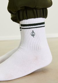 Piedi che indossano calze bianche a coste con strisce nere e un emblema a forma di nota musicale, abbinate a pantaloni verde scuro raccolti alle caviglie.
