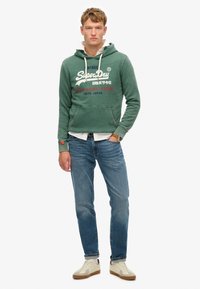 Zielona bluza z kapturem z białymi sznurkami, z nadrukowanym logo i tekstem w czerwieni i niebieskim. W zestawie z niebieskimi dżinsami i beżowymi sneakersami.