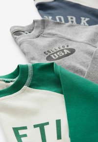 Trois sweat-shirts pliés en blanc-vert, gris et bleu avec des motifs de texte incluant "COLLEGE USA" et "YORK" visibles sur une surface blanche.