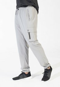 Lichtgrijze joggers met een elastische tailleband, zijzakken en cargozakken met ritsen. Het materiaal lijkt soepel en lichtgewicht.
