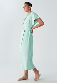 Mėtų žalios spalvos, kelio ilgio suknelė su trumpomis rankovėmis, mygtukų uždarymu, dviem didelėmis kišenėmis ir diržu. Dėvima su šlepetėmis su dirželiais.