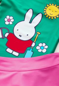 Maillot de bain vert avec un lapin en cartoon portant une chemise rouge et tenant une pelle, entouré de fleurs et d'un soleil. Bas rose avec des détails de couture.