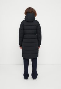 Μαύρο παλτό τύπου puffer με κουκούλα, με ραμμένο σχέδιο και μακριά μανίκια, που φτάνει μέχρι το γόνατο. Φορεμένο πάνω από σκούρο τζιν.