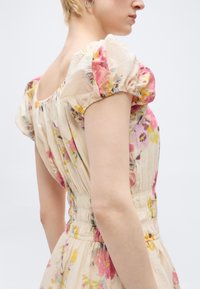 Κοντινό πλάνο ενός ατόμου που φοράει φόρεμα εκρού με λουλουδάτο μοτίβο, φουσκωτά μανίκια και ελαστική μέση, παρουσιάζοντας το πλάι και την πίσω όψη.