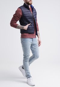 Námořnická puffer vest se zvýšeným límcem přes červený svetr, doplněná světle modrými slim fit džíny a bílými teniskami. Obsahuje zip a prošívaný design.