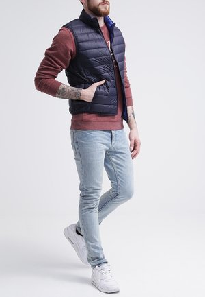 Gilet rembourré bleu marine avec col haut sur un sweater rouge, associé à un jean slim bleu clair et des baskets blanches. Présente une fermeture éclair et un design cousu.