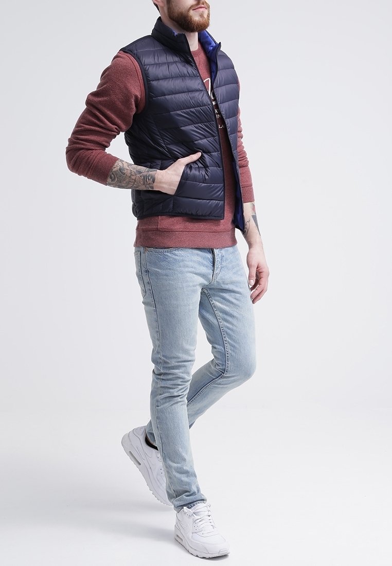 Námořnická puffer vest se zvýšeným límcem přes červený svetr, doplněná světle modrými slim fit džíny a bílými teniskami. Obsahuje zip a prošívaný design.