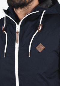 Man in een marineblauwe hoodiejack met witte rits en trekkoorden, met bruine leren patches en een metalen ritslip.