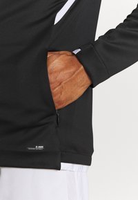 Svart sportsoverall med en dragkedjeficka på sidan, med en slät tygstruktur och vita detaljer längs axeln.