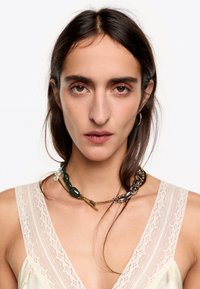 Collar grueso con cuentas verdes, cadena dorada y acentos plateados. Lleva puesto un top de tela ligera con borde de encaje. El cabello castaño cae suelto.