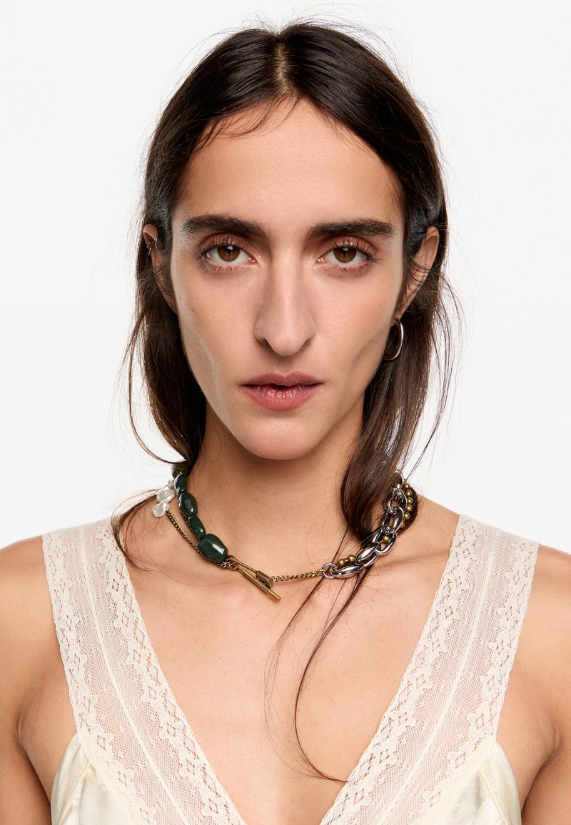 Collar grueso con cuentas verdes, cadena dorada y acentos plateados. Lleva puesto un top de tela ligera con borde de encaje. El cabello castaño cae suelto.