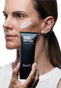 Un tubo nero di "Maschera Notturna Riparativa al Retinolo" tenuto in una mano, con testo bianco; il modello ha una crema applicata sulla guancia.