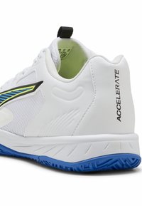 Zapatilla deportiva blanca con suela azul, lengüeta negra, texto "ACCELERATE" en el talón y un diseño a rayas azules y amarillas en el lateral.