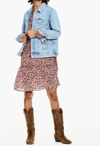 Giacca di jeans blu chiaro su un vestito floreale rosa, con orlo a strati. Stivali cowboy marroni con cuciture decorative.