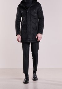 Czarny płaszcz o zamszowej fakturze z kapturem, wyposażony w kieszenie na piersi na suwak, zapięcie na zatrzaski oraz kontrastowe przeszycia. Noszony na czarnych spodniach i butach.