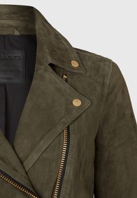 Zielonooliwkowa zamszowa kurtka z klapami, złotymi elementami wykończenia, ukośnym zapięciem na zamek oraz gładką fakturą.